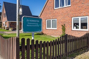 Ein Schild, das ein Objekt zur Miete anzeigt, mit einem Ziegelhaus und einem Holzzaun im Hintergrund.