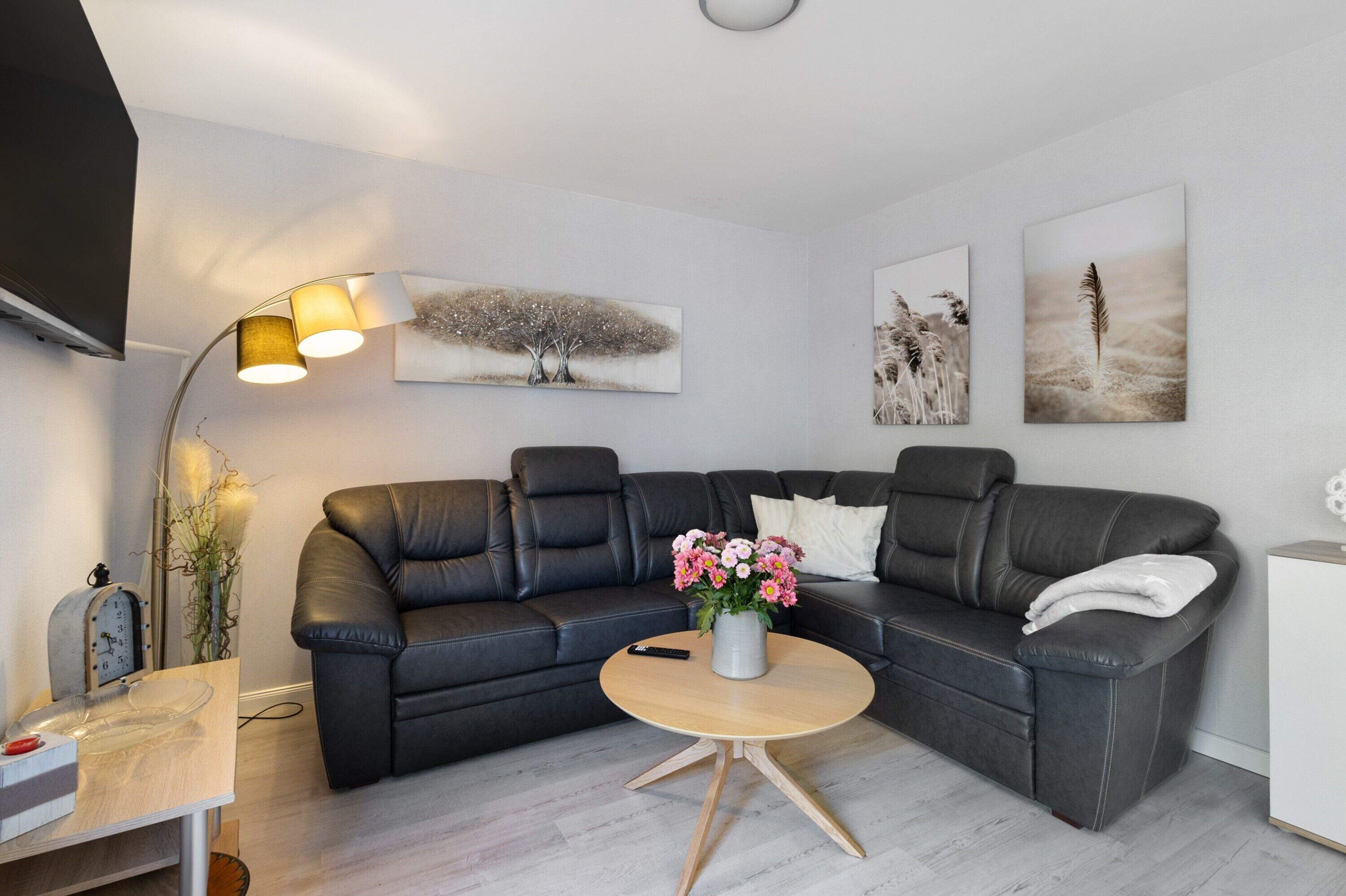 A modern living room with a black leather sofa, a round wooden coffee table with a vase of flowers, a floor lamp, and artwork on the walls.
