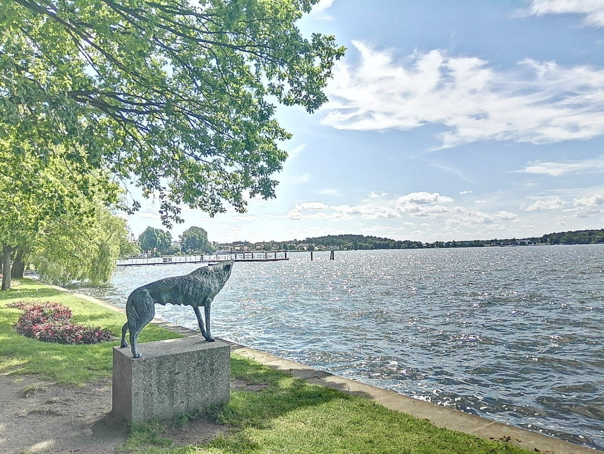 A serene lakeside view with a statue of a wolf on a concrete pedestal, surrounded by lush greenery and a clear sky.