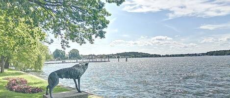A serene lakeside view with a statue of a wolf on a concrete pedestal, surrounded by lush greenery and a clear sky.