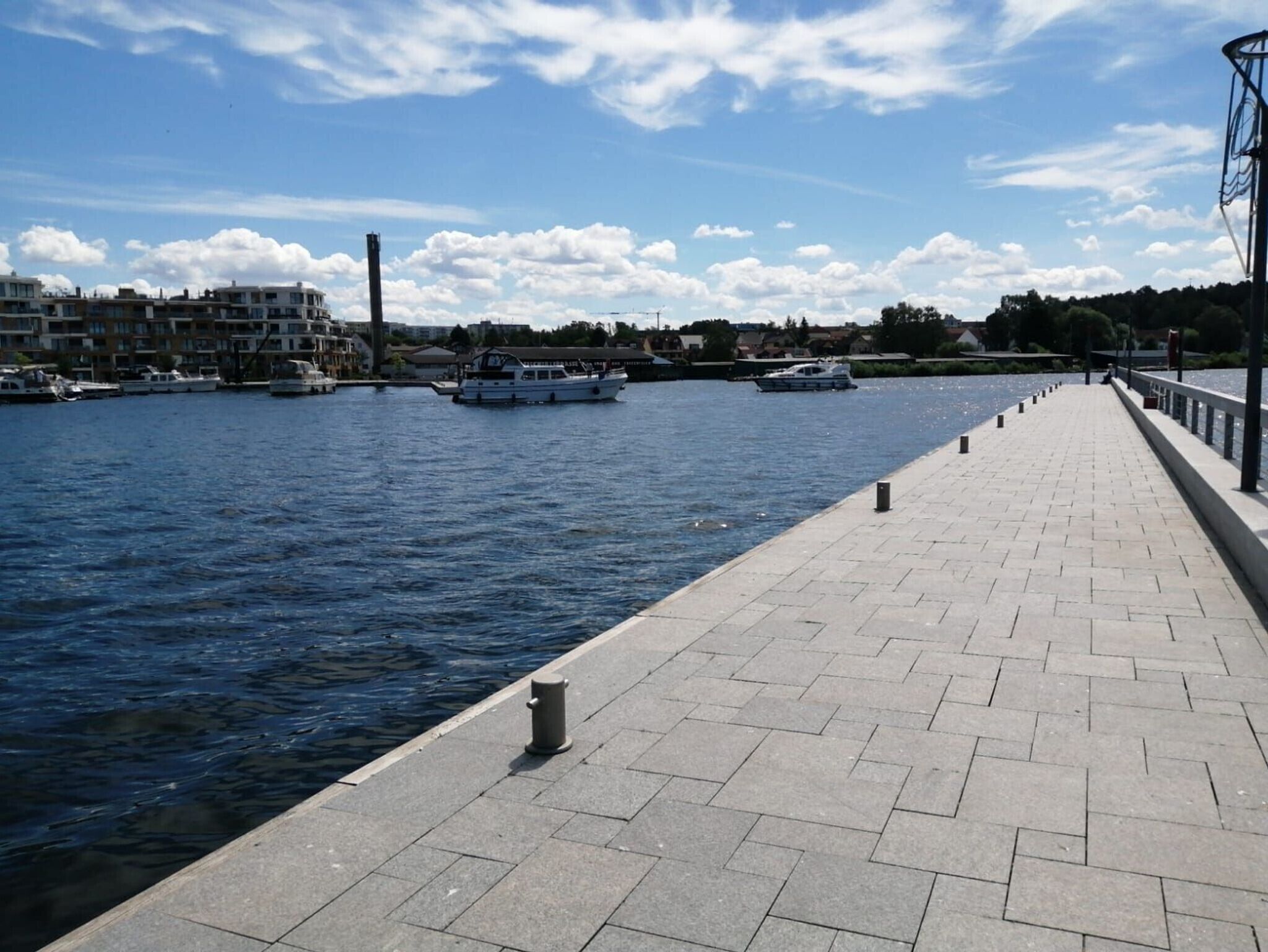 A waterfront promenade with a paved walkway, boats on the water, and buildings in the background.