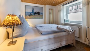 A bedroom with a wooden ceiling, a bed with a striped blanket, a lamp with a textured shade, a painting of a lighthouse, and a window with curtains. - Apartment 'Doppelhaushälfte Ida' with Private Terrace, Shared Garden and Wi-Fi (Meldorf)