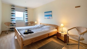 Une chambre à coucher avec un lit en bois, une chaise, une table de chevet avec une lampe, une fenêtre avec des rideaux et un tableau accroché au mur.