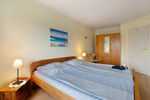 A single bed with a patterned bedspread, a bedside lamp, a wooden nightstand, a wooden door, and a wall-mounted picture.