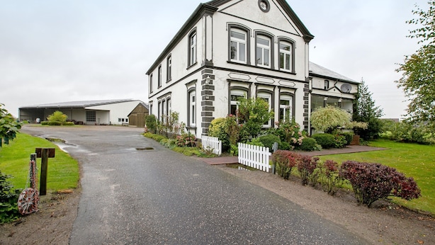 Une maison à deux étages avec une allée menant à l’entrée, entourée d’une clôture en piquets blancs et de divers arbustes.