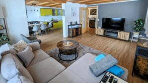 Un salon moderne avec un canapé, une table basse, un téléviseur et une cuisine en arrière-plan.