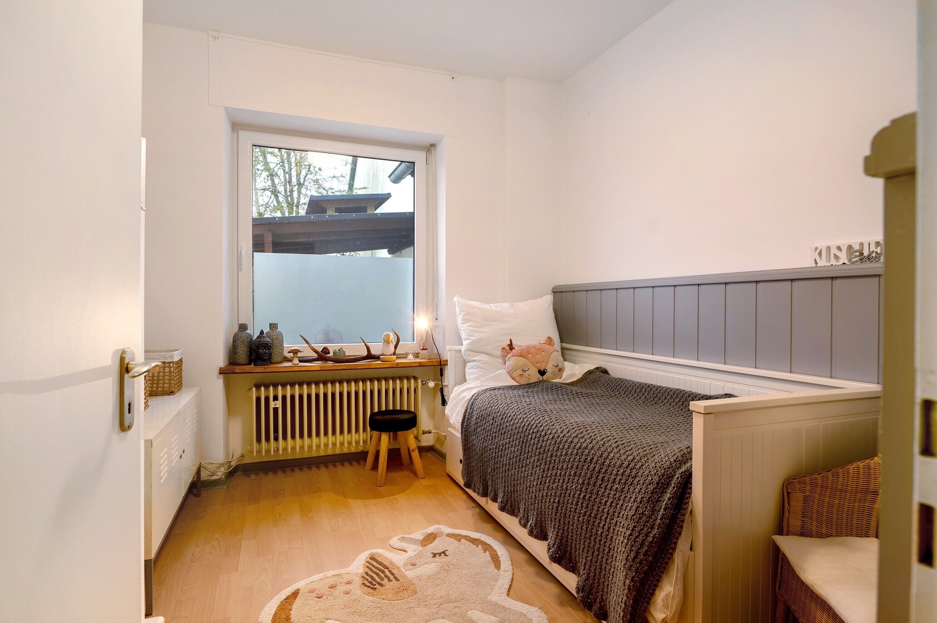 Un dormitorio con cama, radiador, ventana y un taburete pequeño.
