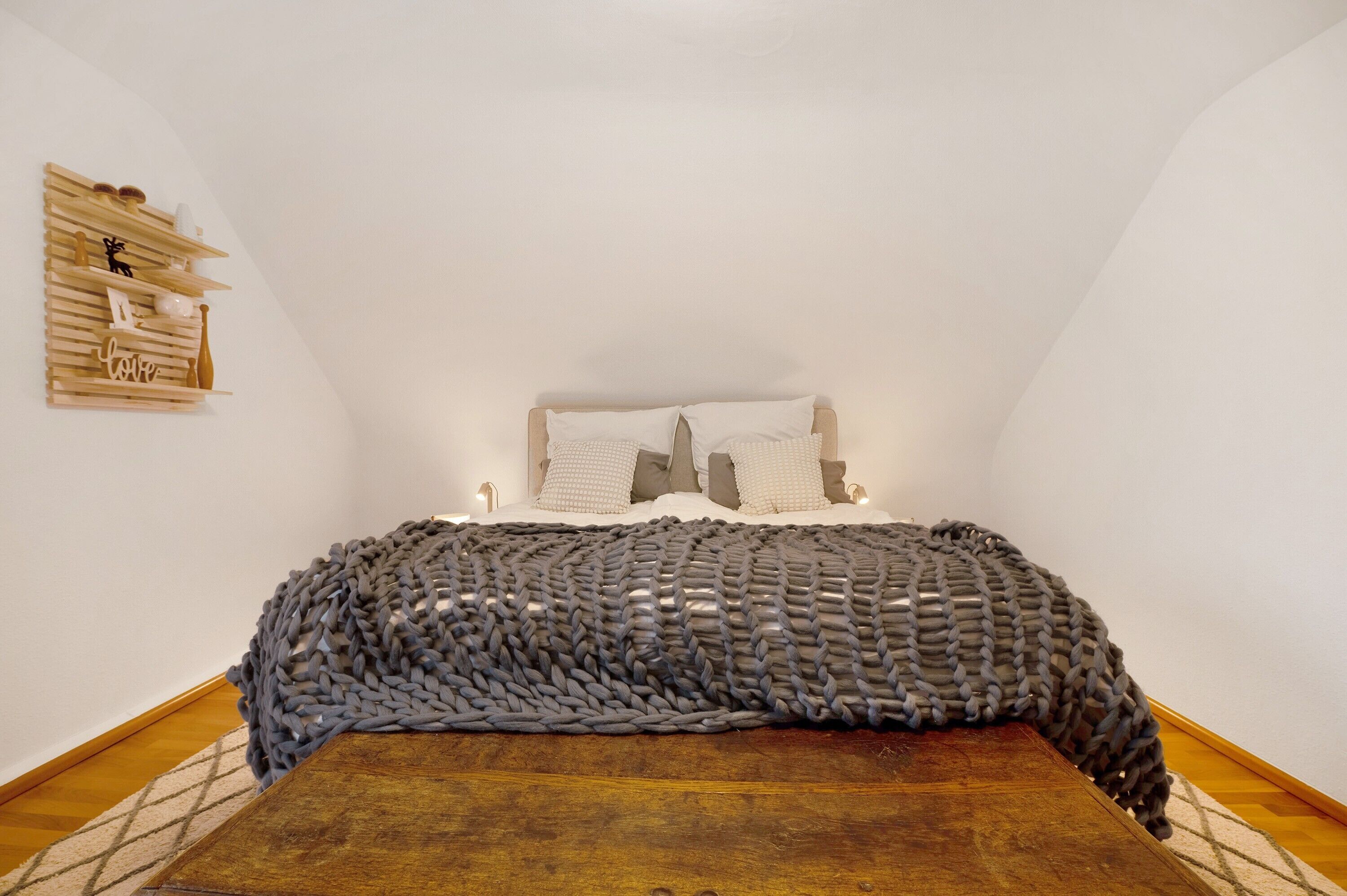 Een slaapkamer met een houten hoofdbord, een grijze gebreide deken en een wandplank met decoratieve objecten.