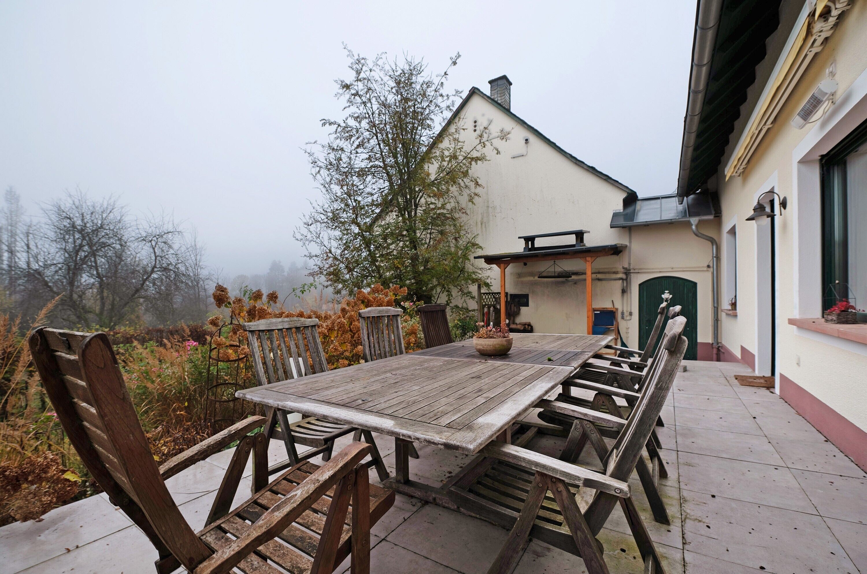 Ein Gartentisch aus Holz mit Stühlen, einer Topfpflanze und einem Gebäude im Hintergrund.