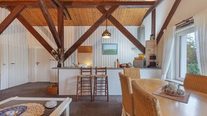 Un interior espacioso con techo alto, vigas de madera, una zona de cocina con taburetes de bar y una mesa de comedor con sillas de mimbre.