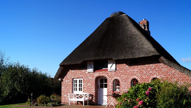Une maison traditionnelle en briques rouges, avec un toit de chaume, un banc blanc et un jardin parsemé de fleurs roses.