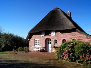 Une maison traditionnelle en briques rouges, avec un toit de chaume, un banc blanc et un jardin parsemé de fleurs roses.
