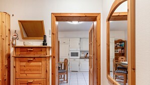 Ett träskåp med lådor, en spegel och ett litet bord med en stol, placerat i en hall som leder till ett kök med vita skåp och en matplats.