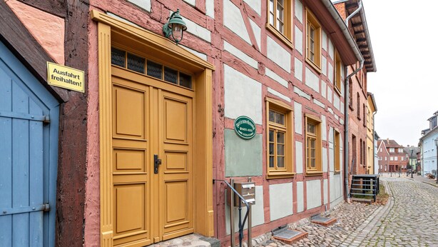 En traditionell byggnad med korsvirkesfasad, en stängd trädörr, en skylt som visar att det finns ett rum att hyra och ett litet utomhussteg som leder till entrén.