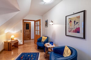 Una habitación con piso de madera, una silla azul, una mesita con bebidas, una lámpara y un cuadro en la pared.