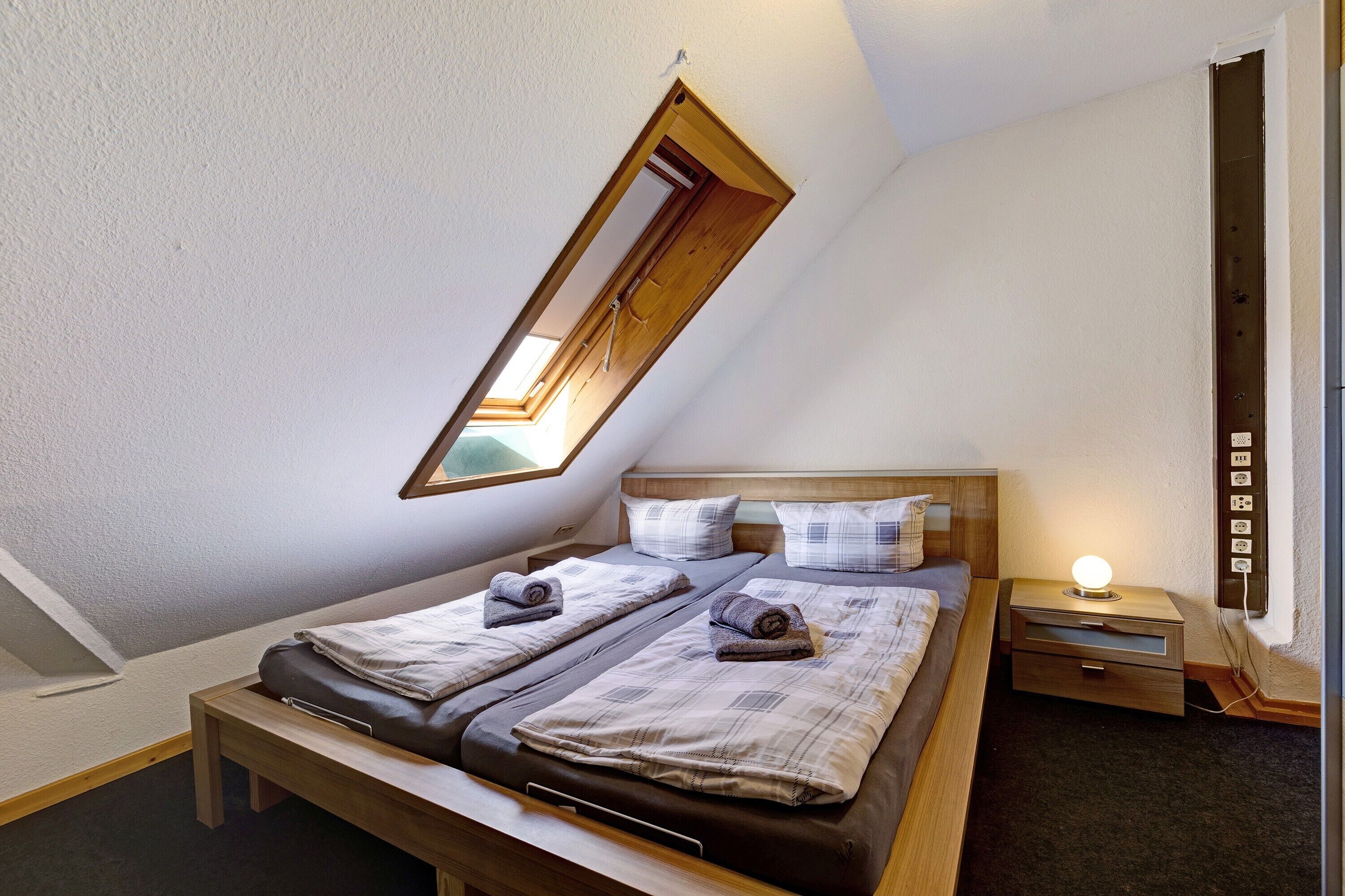 Une pièce avec un plafond en pente, un lit avec deux oreillers, une table de chevet avec une lampe et une prise électrique au mur.
