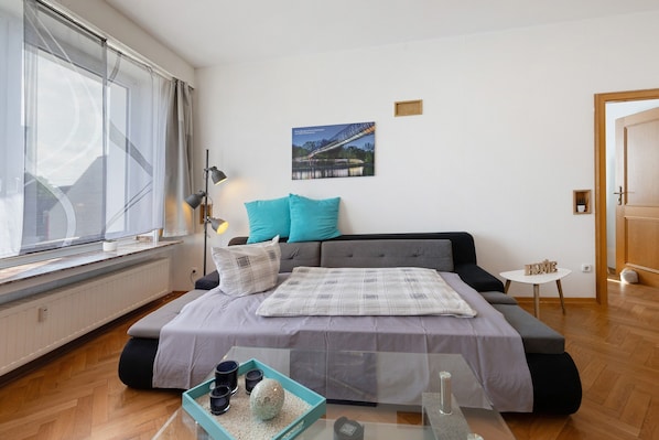 A modern bedroom with a bed, a small table, a window with curtains, a wall-mounted picture, and a door.