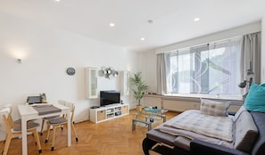 A modern living room with a sofa, a TV, a dining table with chairs, a bookshelf, and a large window with curtains.