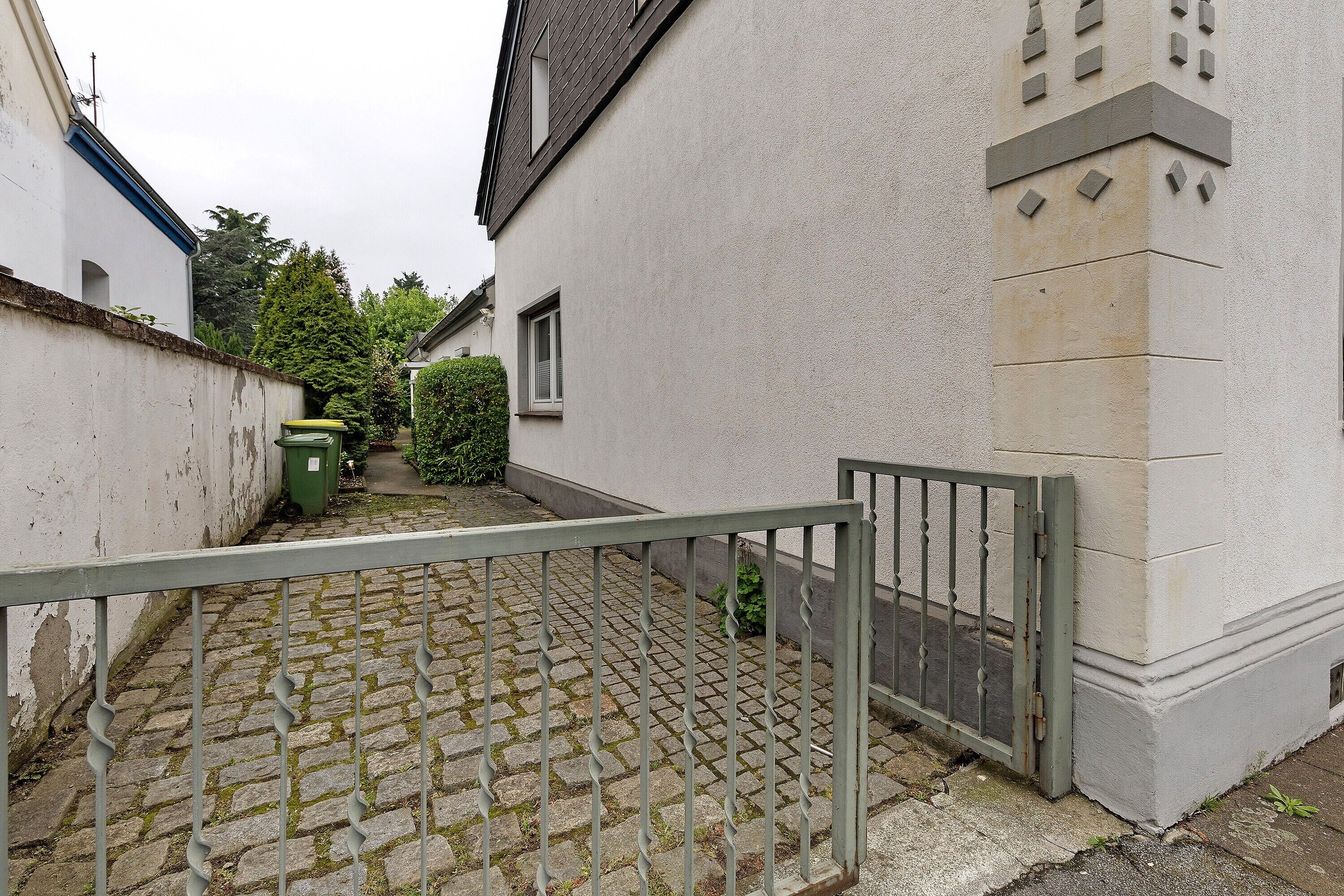 Un étroit sentier pavé de pierres menant à une entrée fermée par un portail, donnant sur un bâtiment à la façade blanche et doté d’une petite fenêtre.
