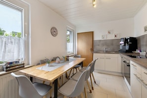 A modern kitchen with a wooden table, grey chairs, a coffee machine, and a window with curtains.