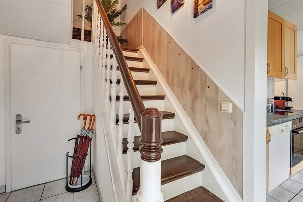 Una escalera con escalones de madera y balaustradas blancas, una puerta con manija, y un portal para paraguas en la pared, con paraguas.