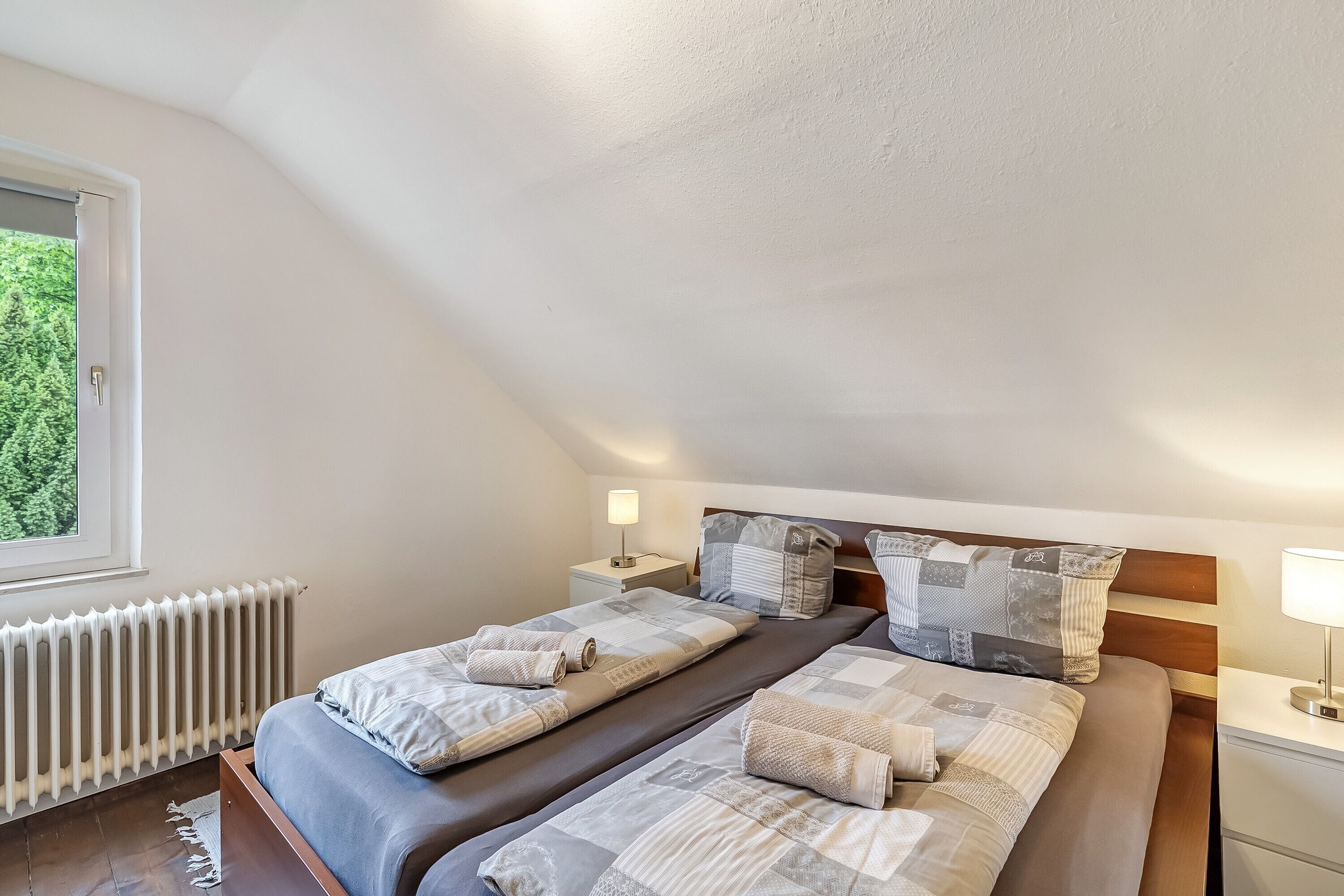 Ein Schlafzimmer mit zwei Betten, einem Heizkörper, Nachttischlampen und einem Fenster mit Blick auf Grünflächen.