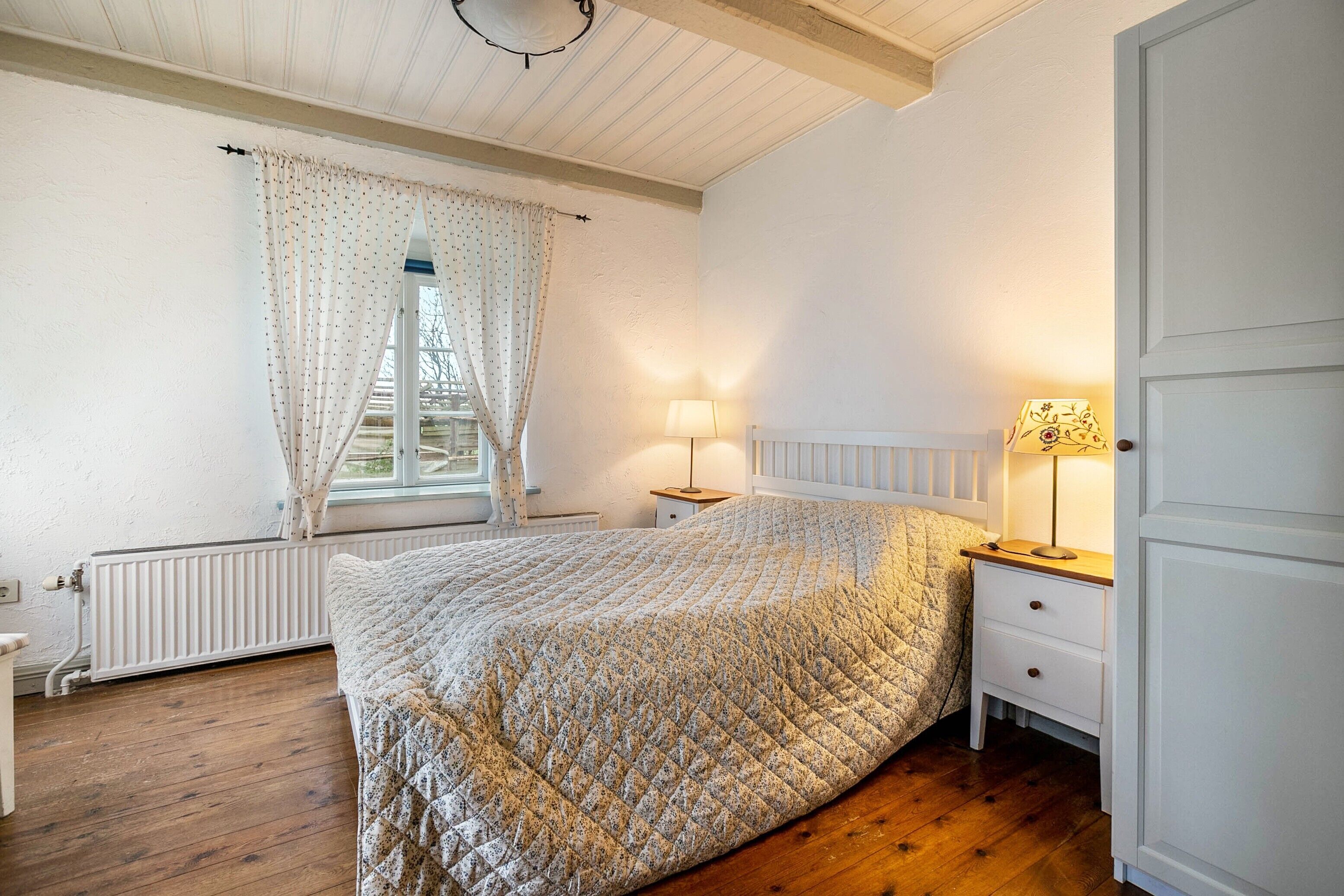 A bedroom with a bed, a nightstand, a lamp, a window with curtains, and a radiator.