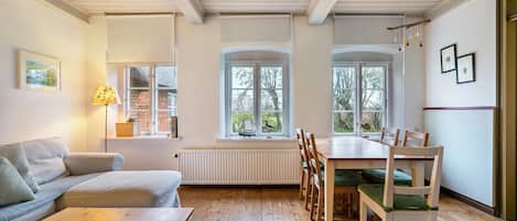 A cozy room with a wooden floor, a sofa, a dining table with chairs, and a window with a view of trees.