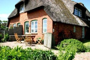 Une maison à toit de chaume, aux murs en briques, avec une table en bois et des chaises, et un jardin à l’avant.