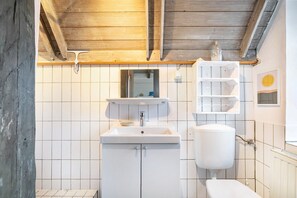 Une salle de bain avec un plafond en bois, des carreaux blancs, un lavabo, des toilettes et une étagère contenant des articles de toilette.