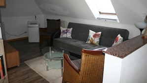 A loft-style living room with a sloped ceiling, a sofa, a wicker chair, a glass coffee table, and a small kitchenette.