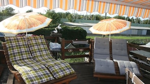 A wooden deck with two lounge chairs and two umbrellas, surrounded by greenery and a building in the background.