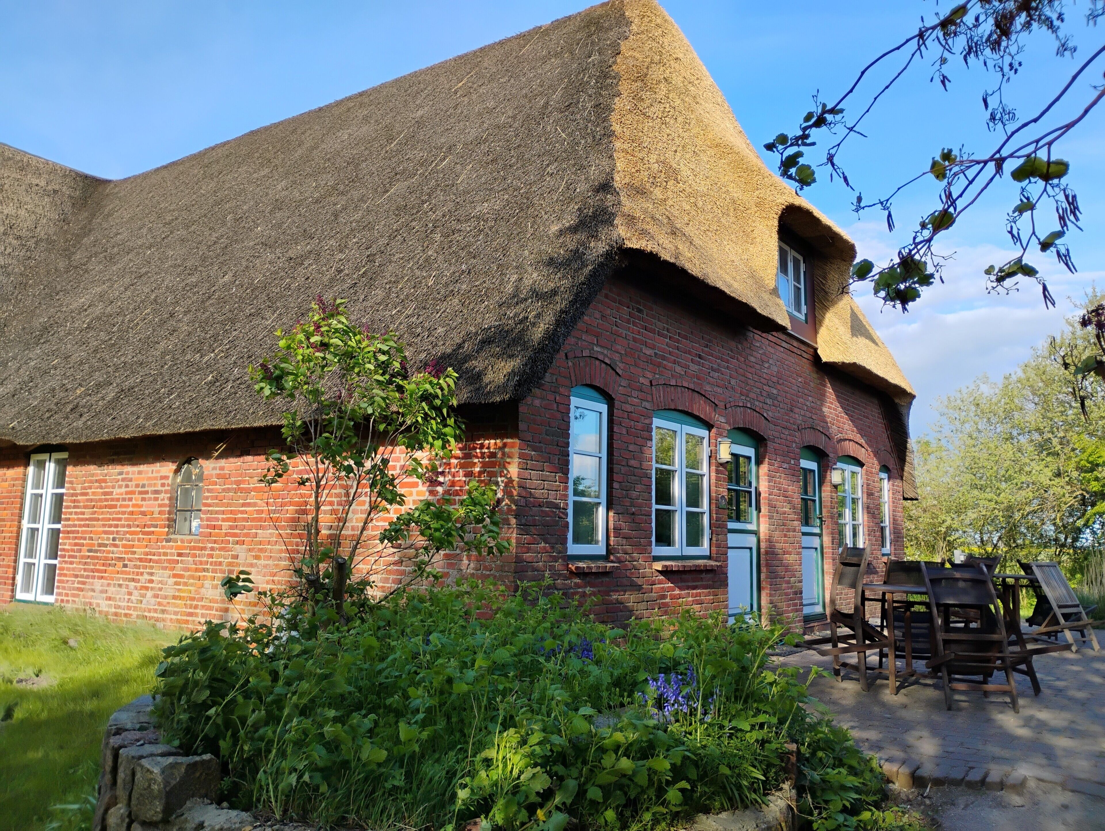 A traditional brick house with a thatched roof, a stone wall, and a garden with various plants and flowers.