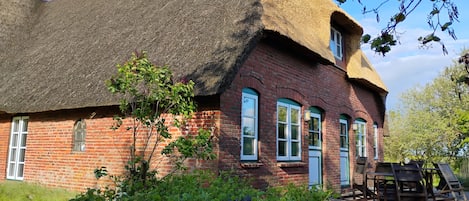 A traditional brick house with a thatched roof, a stone wall, and a garden with various plants and flowers.