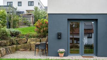 Un bâtiment moderne à la façade grise, doté d’une porte avec des panneaux de verre et d’un petit espace extérieur aménagé avec une table et des chaises.