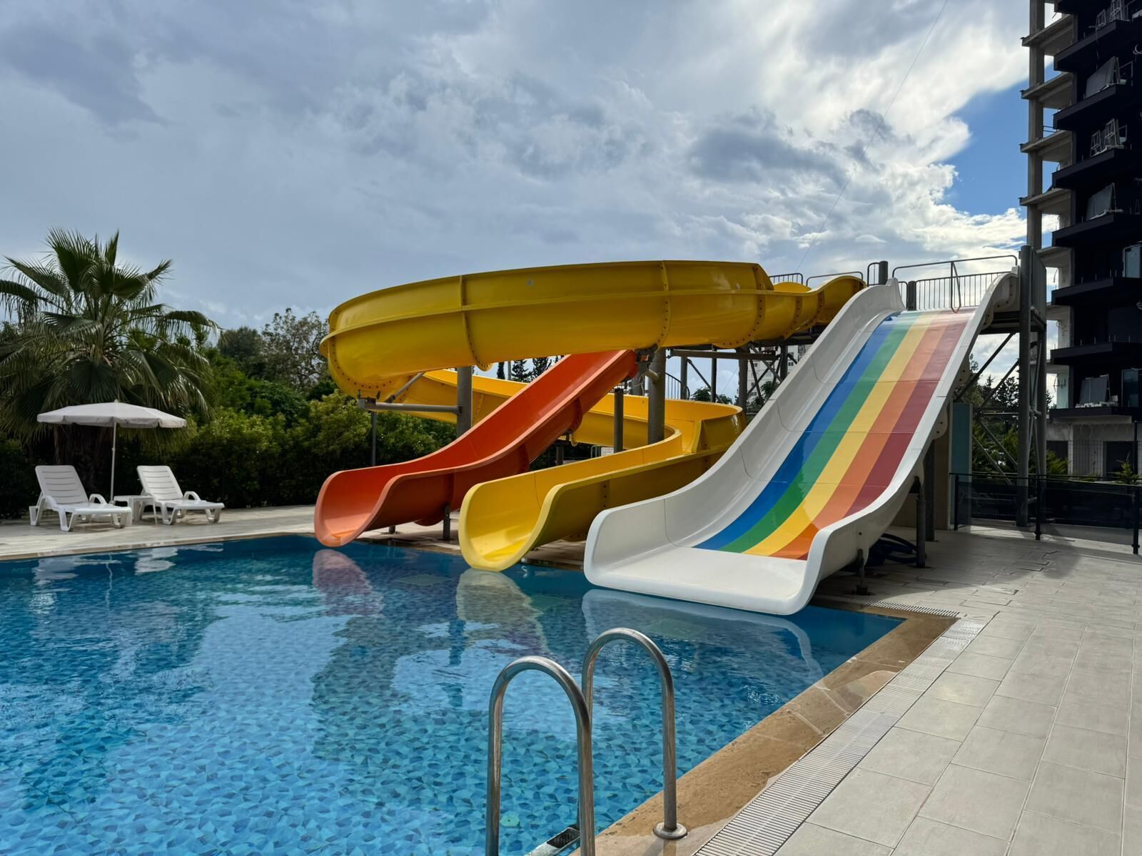 A swimming pool with multiple slides, a lounge area with chairs, and a building in the background.