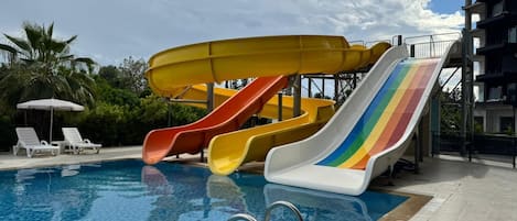 A swimming pool with multiple slides, a lounge area with chairs, and a building in the background.