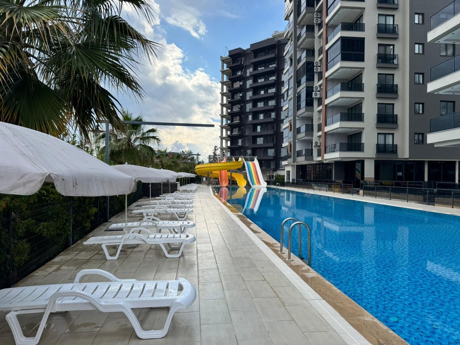 Una piscina con sillas de descanso y sombrillas, un tobogán acuático y modernos edificios de apartamentos al fondo.