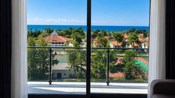 A room with a large window overlooking a coastal area, a balcony with a railing, and a view of the sea and buildings. - Mersin Panorama Otel (Erdemli)