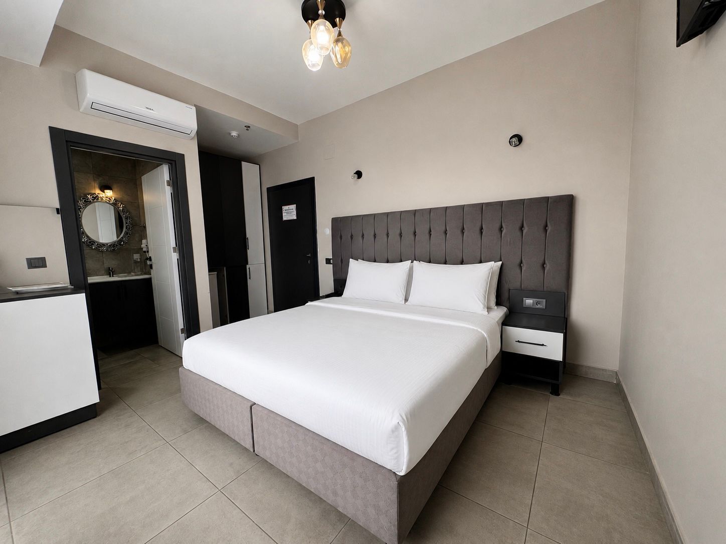 Una habitación de hotel moderna con una cama grande, mesitas de noche, un armario empotrado, un espejo y un equipo de aire acondicionado.
