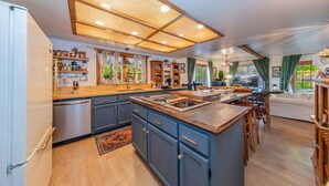 A spacious kitchen with a large island, wooden cabinets, and a built-in oven. There is a dining area with a table and chairs, and a view of the outdoors through large windows. - Sunset Summits Cabin | Hot Tub | Game Room | (Oakhurst)
