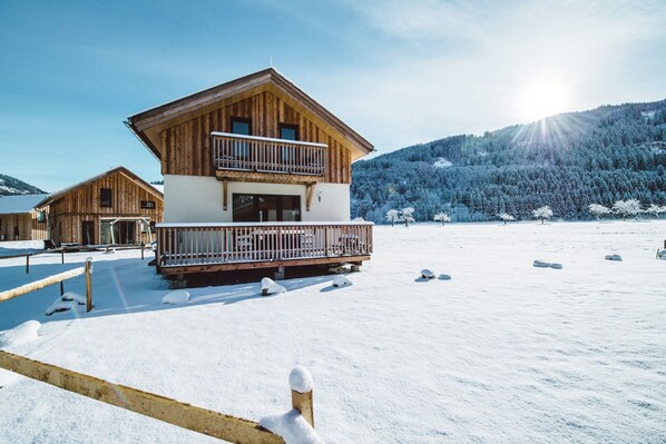 A wooden chalet with a balcony, surrounded by snow-covered ground and trees, with a mountain in the background.