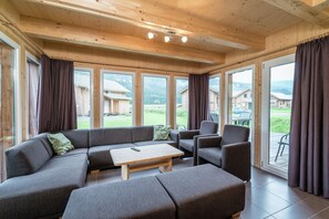 A modern living room with a sectional sofa, a coffee table, and a dining area. Large windows offer a view of a residential area.