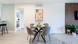 A dining area with a round table, four chairs, and a vase with flowers. There is a painting on the wall above the table and a plant on the right side. - Sleek 1BR: Pool & Fashionable Design (Fort Lauderdale)