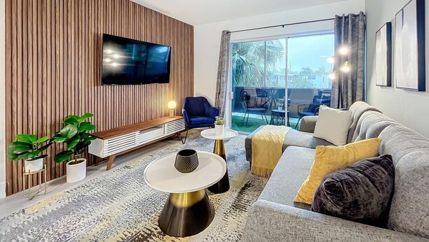 A modern living room with a TV mounted on a wooden wall, a sofa, a blue chair, and a small table with a lamp. - Sleek 1BR: Pool & Fashionable Design (Fort Lauderdale)