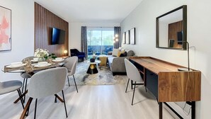 A modern living room with a dining area, a large flat-screen TV, and a large window offering a view of the outdoors. - Sleek 1BR: Pool & Fashionable Design (Fort Lauderdale)