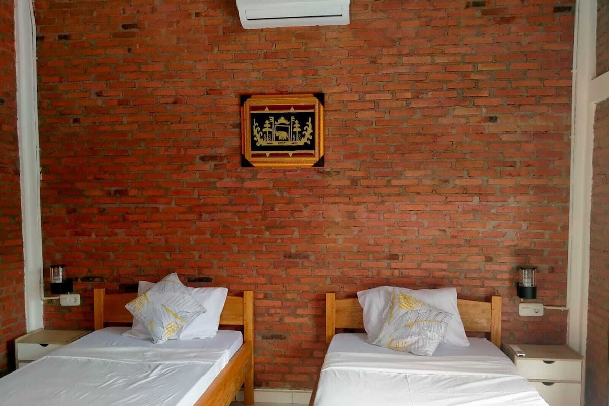 Una habitación con dos camas de madera, ropa de cama blanca, una pared de ladrillo y un cuadro enmarcado.