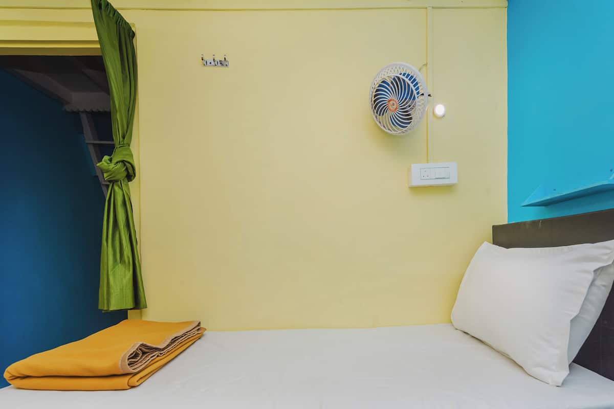 Une pièce avec un lit, un ventilateur fixé au mur, une prise de courant et un rideau vert.