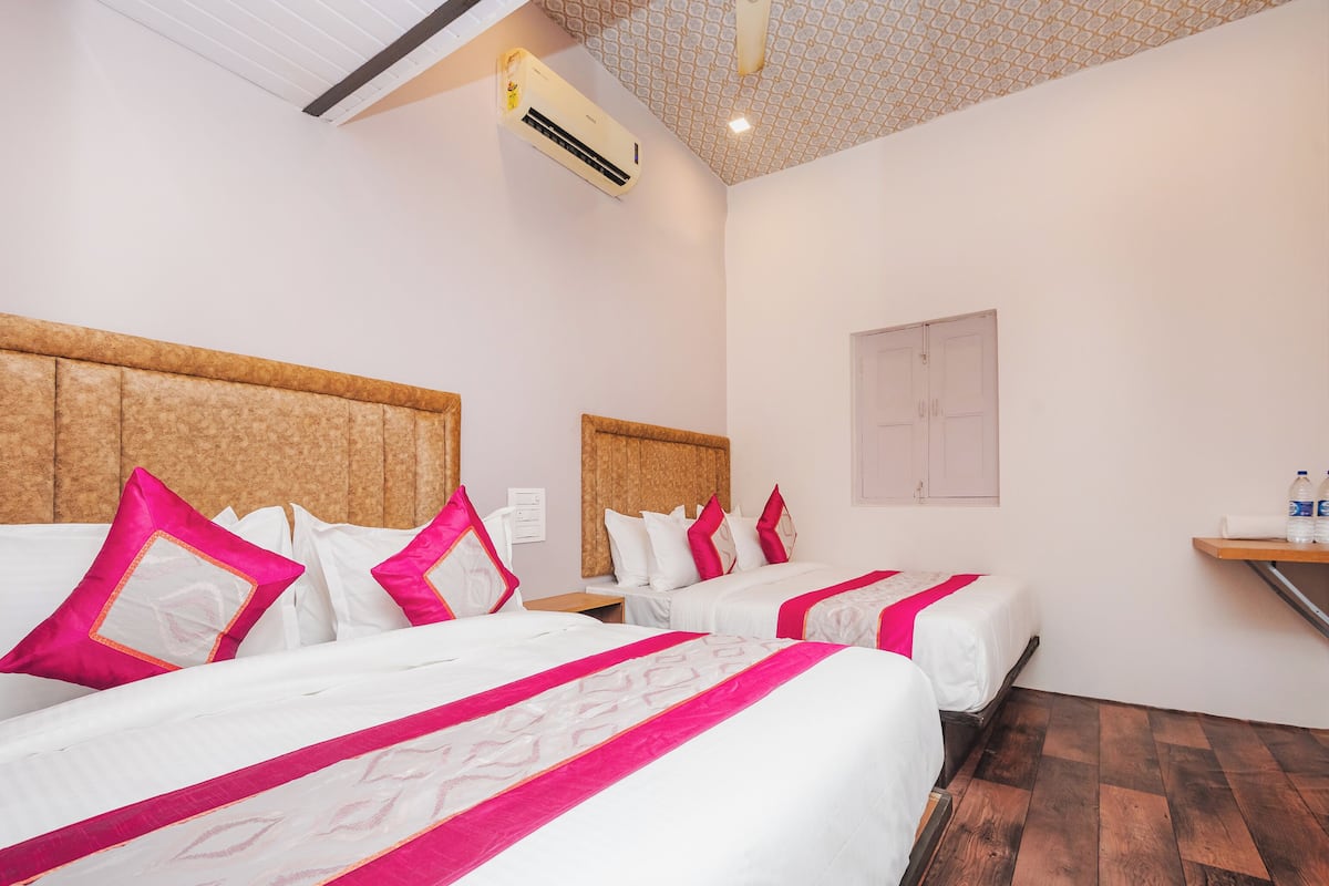 Une chambre d’hôtel avec trois lits, chacun doté de draps blancs et de touches de couleur rose, d’une tête de lit en bois, d’un ventilateur de plafond et d’un distributeur d’eau mural.