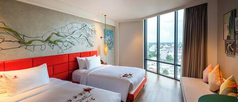 Une chambre d’hôtel avec deux lits, une tête de lit rouge, une grande fenêtre donnant sur la ville et une table verte.